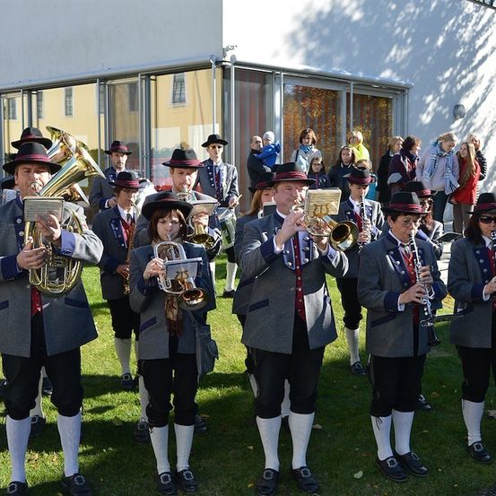 Eine Blaskapelle in traditioneller bayerischer Kleidung spielt auf einem Rasen vor einem Gebäude, während ein Publikum zuschaut.
