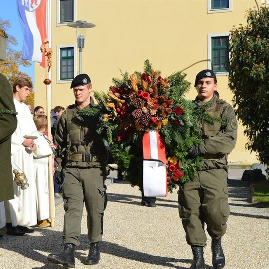 Zwei Soldaten tragen einen Kranz mit Blumen und Tannenzapfen und gehen vor einer Menge her, möglicherweise bei einer Gedenkfeier.