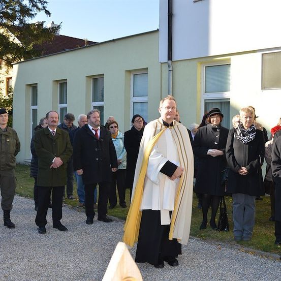 Ein Priester steht vor einer Gruppe von Menschen, möglicherweise während eines Gottesdienstes, mit einem Gebäude im Hintergrund.