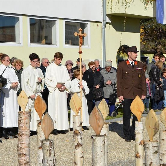 Eine feierliche Zeremonie mit einer Gruppe von Menschen in weißen Gewändern, einem Mann in Uniform und einem Kreuz im Vordergrund. Sie stehen vor einem Gebäude mit Glasfenstern.