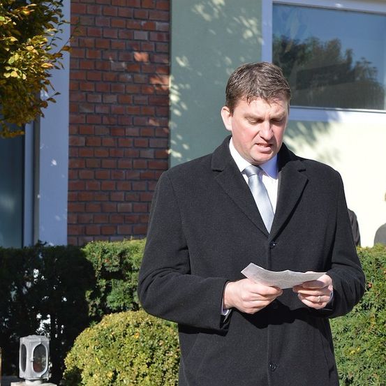 Ein Mann in einem schwarzen Mantel und Krawatte steht vor einem Backsteingebäude und liest ein Stück Papier. Hinter ihm schaut eine Frau mit Brille zu.