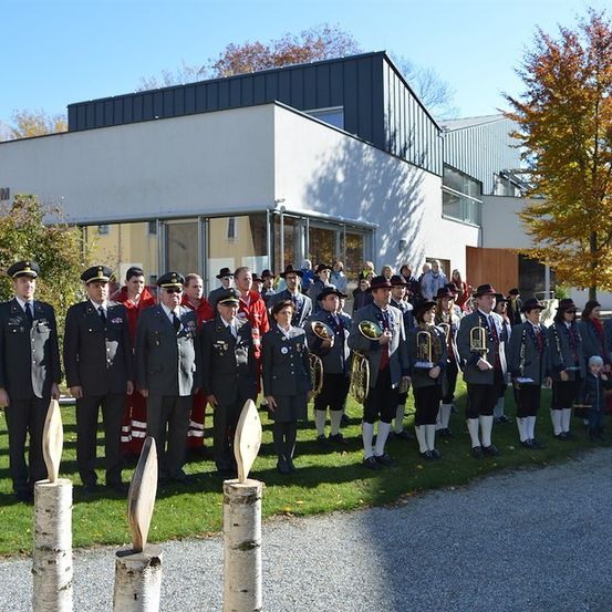 Eine Gruppe uniformierter Personen, möglicherweise eine Militärkapelle, steht vor einem modernen Gebäude an einem sonnigen Tag. Sie halten Blechblasinstrumente und einige tragen Hüte.