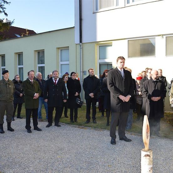 Eine Gruppe von Menschen in formeller Kleidung, darunter einige in Militäruniformen, steht vor einem Gebäude. Ein Mann in einem schwarzen Mantel hält ein Gewehr, während andere hinter ihm stehen.