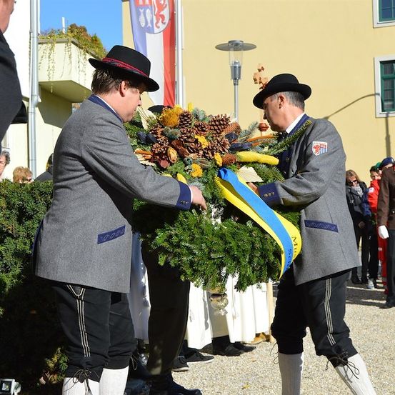 Zwei Männer in traditioneller Kleidung präsentieren einen Kranz mit blauen und gelben Bändern, möglicherweise bei einer feierlichen Veranstaltung. Hinter ihnen ist ein Gebäude mit einer Flagge und Menschen in formeller Kleidung zu sehen.