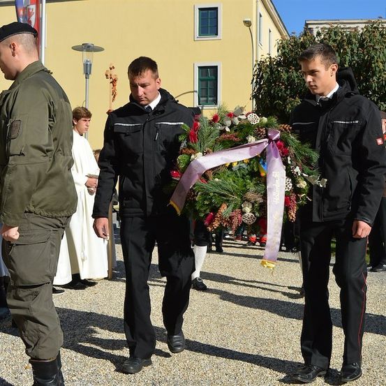 Drei Männer in Uniform, einer hält eine Schleife, gehen an einem Gebäude vorbei, ein Priester im Hintergrund.