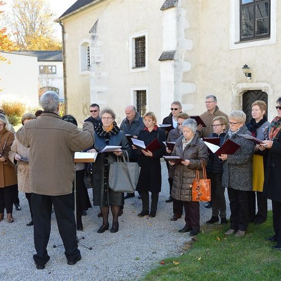 Ein Chor älterer Personen in Winterkleidung steht vor einem Gebäude, hält Liederbücher in den Händen und singt unter der Leitung eines Mannes.