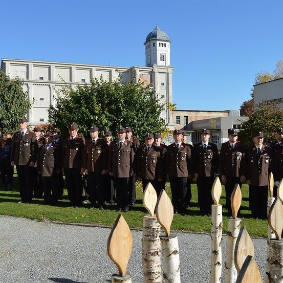 Eine Gruppe uniformierter Personen steht vor einem Gebäude mit einem Turm an einem sonnigen Tag. Sie stehen in Reihen, mit einigen Kerzen vor ihnen.
