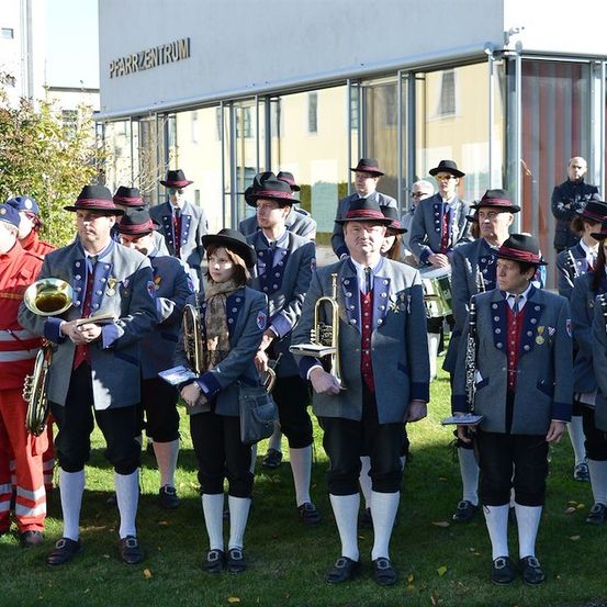 Eine Gruppe von Musikern in traditionellen Uniformen steht vor einem Gebäude mit dem Schild 'Pfarrzentrum'. Sie halten verschiedene Musikinstrumente in der Hand und scheinen sich auf eine Aufführung vorzubereiten.
