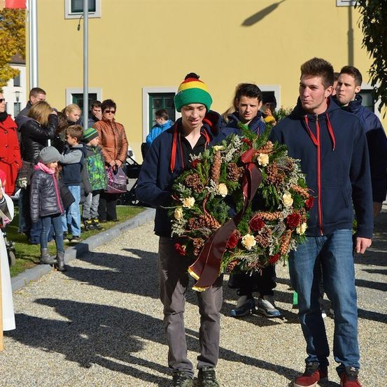 Zwei Männer tragen einen Kranz aus Blumen und Tannenzapfen vor einer Menschenmenge. Der Mann auf der linken Seite trägt einen bunten Hut und eine Jacke. Der Mann auf der rechten Seite trägt eine blaue Jacke und Jeans.