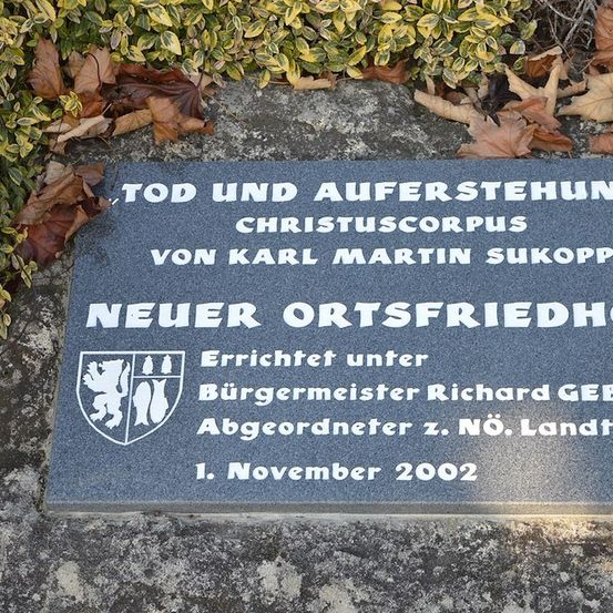 Eine Steinplatte auf einem Betonsockel trägt die Aufschrift 'Tod und Auferstehung Christuscorpus von Karl Martin Sukopp'. Sie wurde unter Bürgermeister Richard Gebe am 1. November 2002 errichtet.