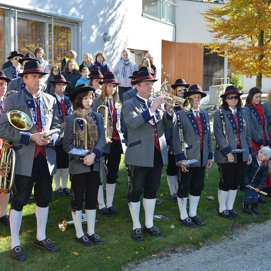Eine Gruppe von Musikern in traditioneller bayerischer Kleidung spielt auf einem Rasenfeld vor einem Gebäude und Bäumen.