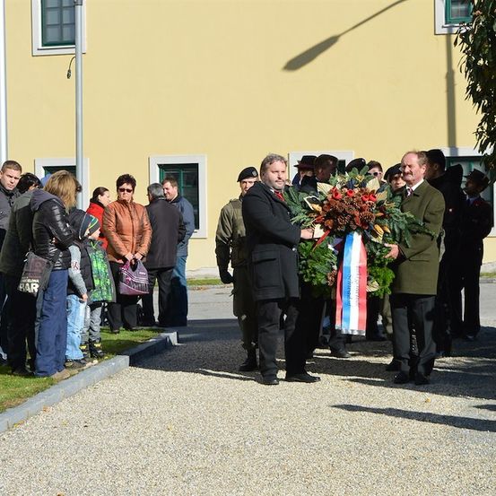 Eine Gruppe von Menschen steht vor einem gelben Gebäude, einige halten Kränze. Zwei Männer in formeller Kleidung legen Kränze auf ein Denkmal.