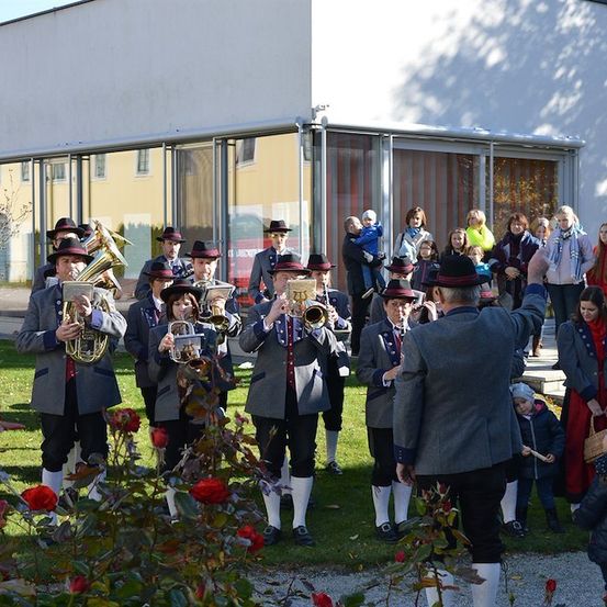Eine Brassband spielt vor einem modernen Gebäude vor einer Menge Zuschauer. Die Bandmitglieder tragen traditionelle Uniformen und spielen verschiedene Instrumente.