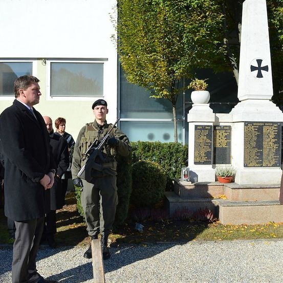 Ein Mann in Uniform steht vor einem Denkmal mit einem Gewehr, während ein Mann in Anzug in der Nähe steht.