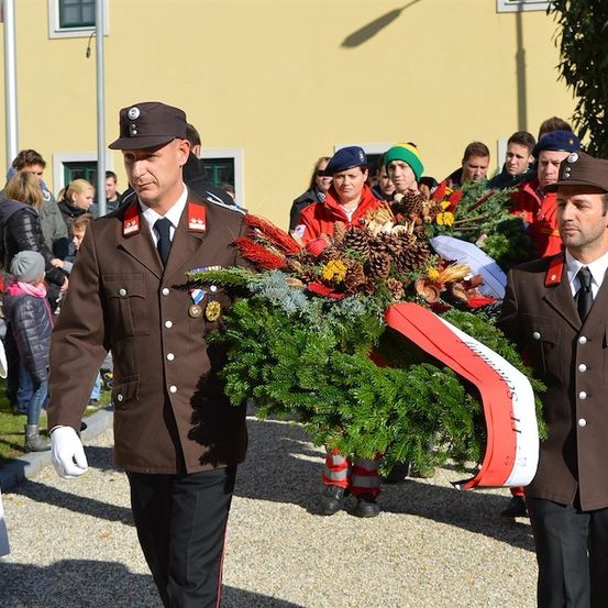 Zwei Männer in Militäruniformen tragen einen Kranz mit Tannenzapfen und roten und weißen Bändern und gehen in einer Prozession vor einem Gebäude.