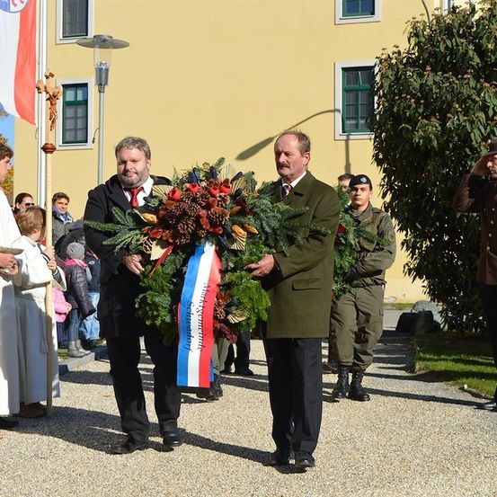 Zwei Männer tragen einen Kranz mit einer Flagge in der Mitte, vor einem Gebäude mit einem Fahnenmast, begleitet von einem Priester und Militärpersonal.