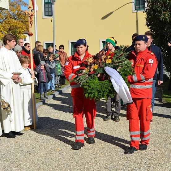 Zwei Feuerwehrleute in roten Uniformen halten einen Kranz mit Blumen und Tannenzapfen und stehen vor einer Gruppe von Menschen, einschließlich eines Priesters in Weiß.
