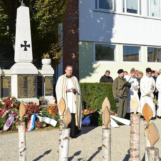 Ein Priester steht vor einem Denkmal mit einem Kreuz, umgeben von Menschen in weißen Gewändern und einem Soldaten mit einem Gewehr, vor einem Gebäude mit Fenstern.