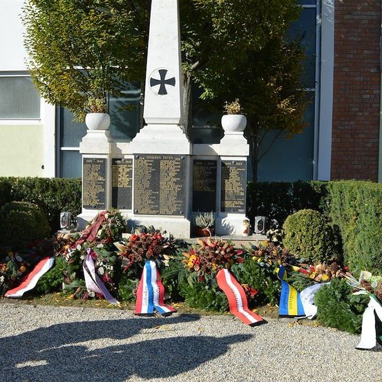 Ein Gedenkmonument mit einem Kreuz und Tafeln mit Namen, umgeben von Kränzen und Flaggen, vor einem Backsteingebäude.