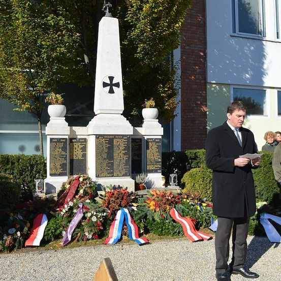 Ein Mann in einem schwarzen Anzug steht vor einem Denkmal mit einem Kreuz oben, hält ein Stück Papier in der Hand. Hinter ihm steht ein Soldat Wache.