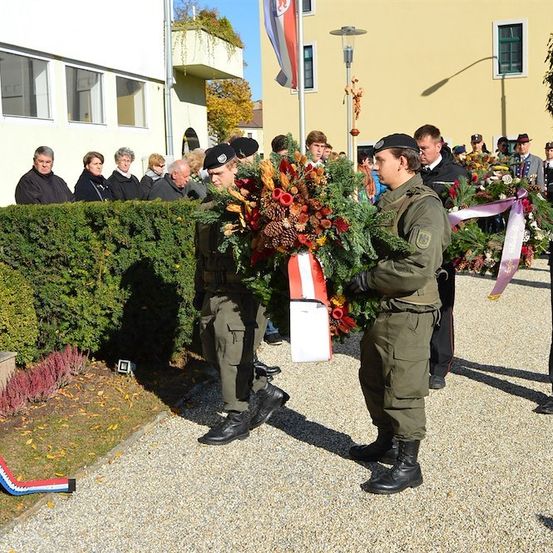 Zwei Soldaten in Uniform halten Kränze mit Blumen und Bändern und stehen vor einem Gebäude, das von Menschen beobachtet wird.