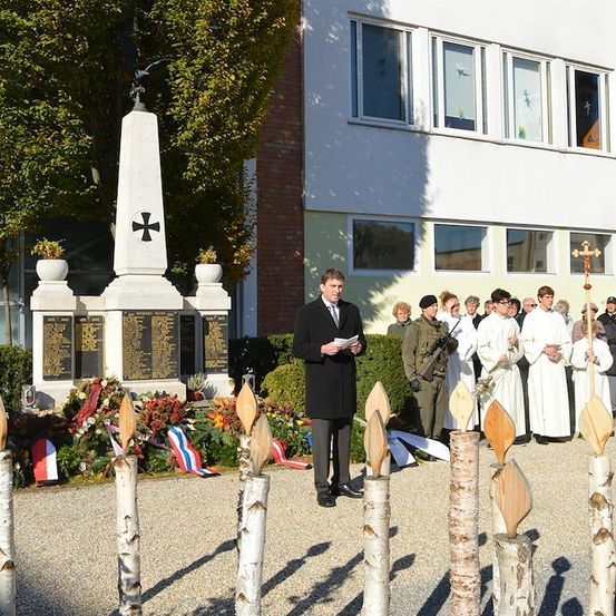 Eine feierliche Versammlung vor einem Denkmal, mit einem Mann, der aus einem Papier liest, umgeben von Menschen in weißen Gewändern und einem Kreuz am Gebäude.