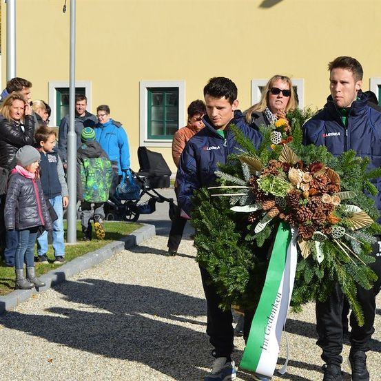 Zwei Männer tragen einen Kranz mit Kiefernzapfen und Grün vor einer Menschenmenge, darunter Kinder und Erwachsene, vor einem gelben Gebäude.