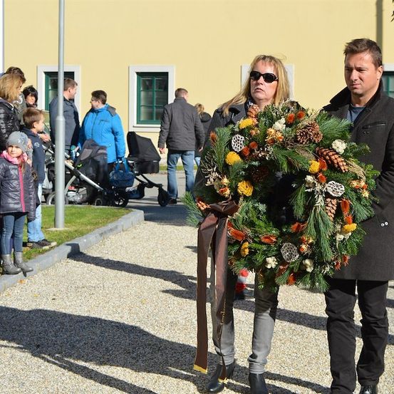 Ein Mann und eine Frau mit einem Kranz gehen an einer Menschenmenge vorbei, darunter Kinder mit Kinderwagen, vor einem gelben Gebäude.