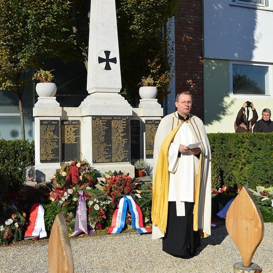 Ein Priester steht vor einem Denkmal mit Kränzen und Flaggen und liest von einem Papier. Dahinter ein Gebäude mit einem Kreuz auf der Spitze und Leute, die Fotos machen.