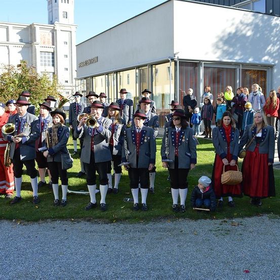 Eine Gruppe von Menschen in traditionellen Kostümen steht auf einem Rasen und führt möglicherweise eine musikalische Veranstaltung durch. Einige von ihnen halten Musikinstrumente. Im Hintergrund befinden sich Gebäude und Bäume.