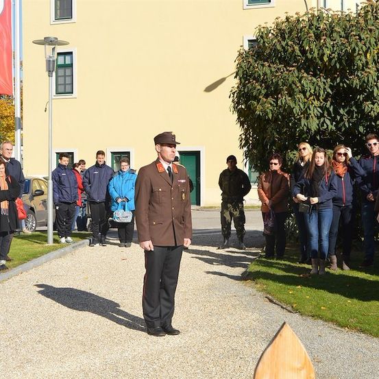 Ein Militäroffizier steht vor einem Gebäude mit einer Flagge, während eine Gruppe von Menschen in verschiedenen Jacken und Stiefeln hinter ihm steht, einige schauen ihn an.