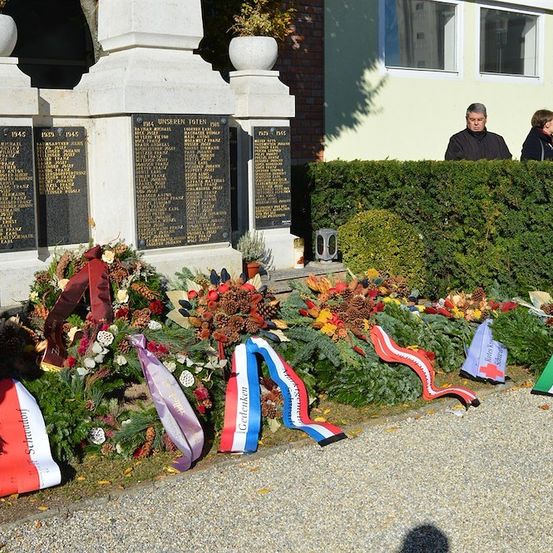 Ein Denkmal mit Kränzen und Bändern vor einem Gebäude, hinter dem Menschen hinter einer Hecke stehen. Das Denkmal hat Tafeln mit Namen und Jahren.