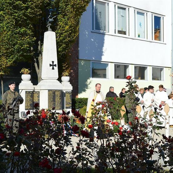 Eine feierliche Zeremonie mit Geistlichen und Militärangehörigen vor einem Denkmal mit roten Rosen und einem weißen Gebäude im Hintergrund.