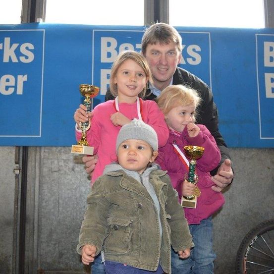 Ein Mann und drei Kinder stehen zusammen und halten Trophäen, wobei zwei Mädchen in rosa Jacken und ein Junge in einer grünen Jacke sind. Sie lächeln und posieren für ein Foto vor einem blauen Hintergrund mit Text.