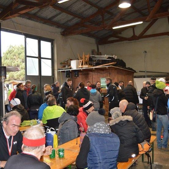 Eine große Gruppe von Menschen in Winterkleidung versammelt sich in einem großen Raum mit Holzbänken und Tischen. Einige sitzen, andere stehen und reden. Es gibt auch einige Leute, die Mützen und Brillen tragen. Der Raum hat eine Holzdecke und eine Glaswand.