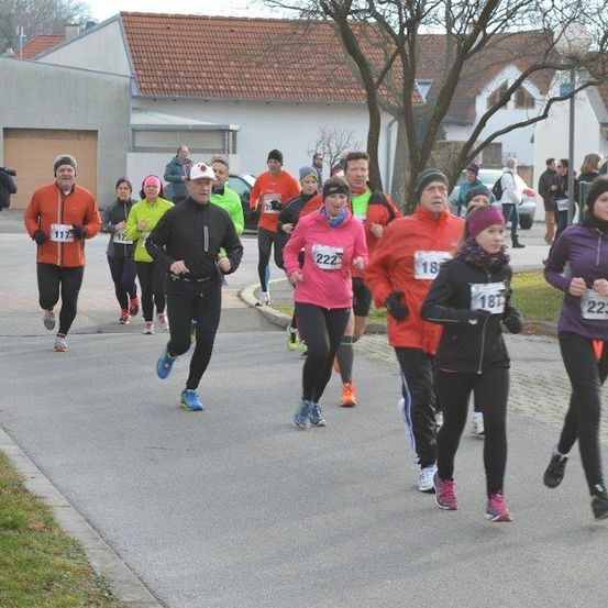 Eine Gruppe von Läufern in verschiedenfarbigen Jacken und Turnschuhen nimmt an einem Rennen auf einer asphaltierten Straße teil. Zuschauer und geparkte Autos sind im Hintergrund zu sehen.