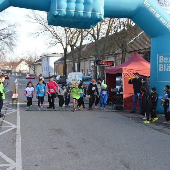 Eine Gruppe von Kindern in Laufausrüstung startet einen Lauf auf einer Straße, mit einem blauen Bogen und einem Zelt in der Nähe.