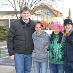 Vier Personen stehen zusammen auf einem Parkplatz und lächeln für ein Foto. Ein Mann hält ein Mikrofon, während die anderen in Winterkleidung gekleidet sind.
