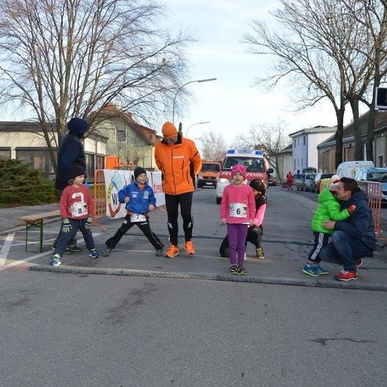 Eine Gruppe von Kindern und Erwachsenen steht an einer Straßenecke, wahrscheinlich bei einem Laufevent. Ein Mann in einer orangefarbenen Jacke geht vor ihnen.