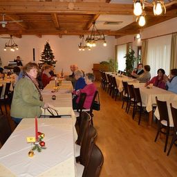 Ein Speisesaal mit langen Tischen, die jeweils mit einer Kerze und Weihnachtsdekoration geschmückt sind, ist mit älteren Menschen gefüllt, die sitzen und sich unterhalten. Der Raum verfügt über Holzböden, Holzbalken und einen Weihnachtsbaum im Hintergrund.