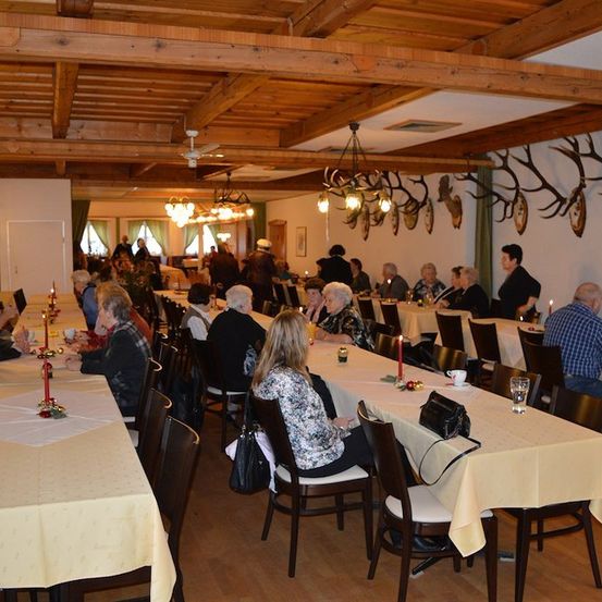 Ein Speisesaal mit langen Tischen, Holzbalken und Geweihen an der Wand. Menschen sitzen an Tischen, einige mit Kerzen.