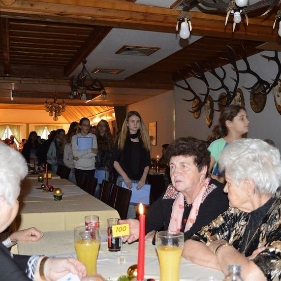 Eine Gruppe älterer Frauen sitzt an einem Tisch mit Kerzen und Getränken. Zwei Frauen schauen sich an, während eine andere Frau in die Kamera schaut. Eine Nummer 100 steht auf einer gelben Karte in der Nähe einer Kerze.