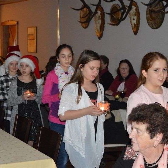 Eine Gruppe junger Mädchen und Frauen hält Kerzen in einem Raum, möglicherweise während einer Weihnachtsveranstaltung. Sie tragen Winterkleidung und einige tragen Weihnachtsmützen. Der Raum hat einen Tisch mit gelber Tischdecke, Stühlen und einer Wand mit Hirschgeweihen.