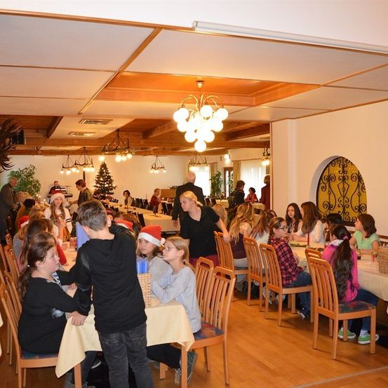 Eine Gruppe von Menschen, einschließlich Kindern, sitzt an Tischen in einem Essbereich, möglicherweise für eine Weihnachtsfeier. Der Raum ist mit Weihnachtsbäumen und Lichtern geschmückt.