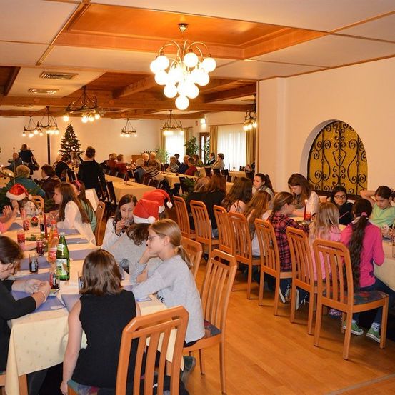 Ein Speisesaal voller Menschen, die an Tischen sitzen, einige mit Weihnachtsmützen. Der Raum hat Holzböden, weiße Wände und einen Weihnachtsbaum in der Ecke.
