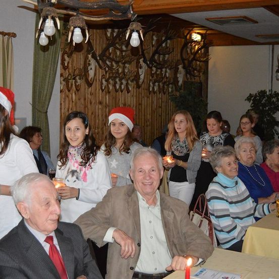 Eine Gruppe von Menschen, darunter ältere Personen und junge Mädchen, versammelt sich in einem Raum und hält Kerzen in den Händen. Sie scheinen einen festlichen Anlass zu feiern.
