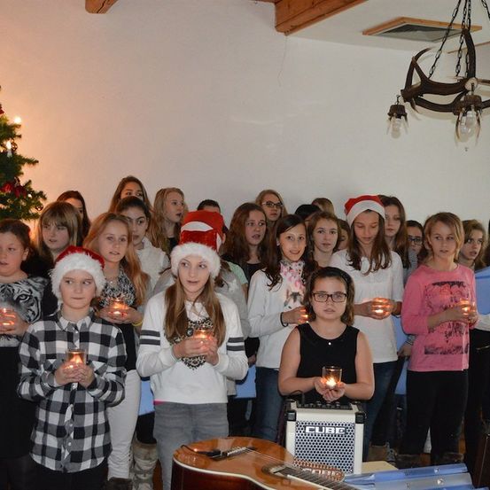 Eine Gruppe von Kindern mit Weihnachtsmützen, die Kerzen in einem Raum mit einem Weihnachtsbaum und einer Gitarre halten.