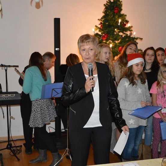 Eine Frau in einer schwarzen Jacke spricht in ein Mikrofon vor einem Weihnachtsbaum, mit einer Gruppe von Mädchen in Weihnachtsmützen und Ordnern hinter ihr.