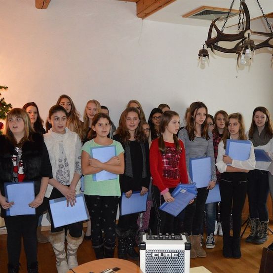 Eine Gruppe junger Mädchen, in Winterkleidung gekleidet, steht in einem Raum und hält blaue Ordner. Ein Weihnachtsbaum und eine Kronleuchter sind im Hintergrund zu sehen.