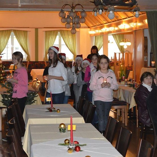 Eine Gruppe von Kindern mit Weihnachtsmützen steht in einem Esszimmer und hält Kerzen. Der Raum ist mit Weihnachtsdekorationen geschmückt, darunter ein Baum und Kerzen auf den Tischen.